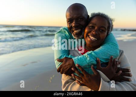 Couple senior afro-américain profite d'une étreinte au coucher du soleil sur la plage. Leur moment joyeux capture un cadre extérieur romantique. Banque D'Images
