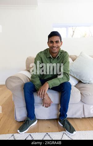 Jeune homme biracial sourit assis sur un canapé à la maison. Sa pose décontractée et son intérieur lumineux créent une atmosphère détendue et accueillante inaltérée. Banque D'Images