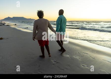 Couple senior afro-américain profite d'une promenade sur la plage au coucher du soleil. Leur promenade sereine le long du rivage met en évidence un moment de compagnie paisible. Banque D'Images