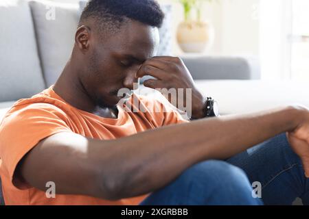 Homme afro-américain réfléchi assis sur le sol dans le salon ensoleillé tenant le nez. Santé mentale, dépression, solitude, émotion, vie domestique et Banque D'Images