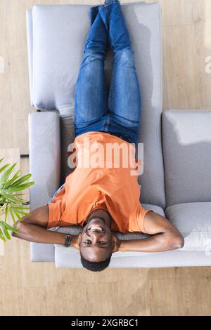 Portrait aérien de l'homme afro-américain heureux allongé sur le canapé dans le salon ensoleillé souriant. Détente, bien-être, vie domestique et style de vie, unalt Banque D'Images