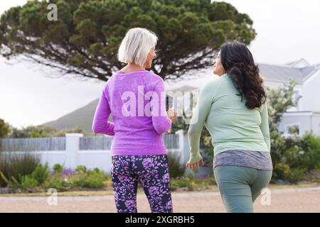 Vue arrière de deux amies féminines âgées heureuses diverses marchant pour l'exercice dans la route de campagne, parlant. Amitié, forme physique, exercice, activité et santé Banque D'Images