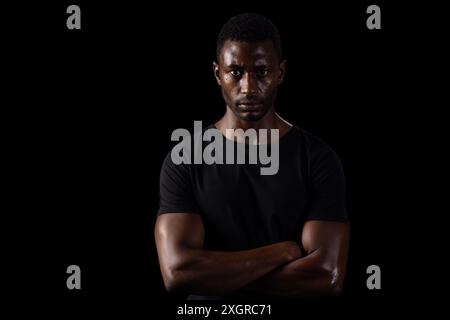 Homme afro-américain se tient confiant sur un fond sombre sur fond noir. Son regard intense et ses bras croisés suggèrent détermination et str Banque D'Images
