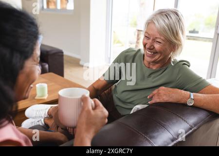 Deux amies âgées heureuses diverses prenant un café et parlant assis sur le canapé dans le salon. Amitié, détente, retraite, vie domestique et Banque D'Images