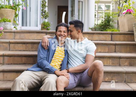 Couple gay diversifié profite d'un moment à l'extérieur, inchangé. Ils partagent une étreinte chaleureuse sur les marches de leur maison. Banque D'Images