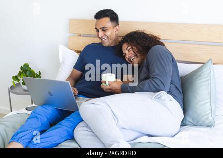 Couple biracial gay aime le temps de loisirs dans le lit, avec espace de copie. Ils partagent un moment agréable à la maison avec un ordinateur portable et un café. Banque D'Images