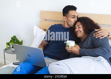 L'homme du moyen-Orient embrasse une jeune femme biraciale dans un cadre de chambre confortable. Ils partagent un moment de détente avec un ordinateur portable et un café, symbolisant le h moderne Banque D'Images