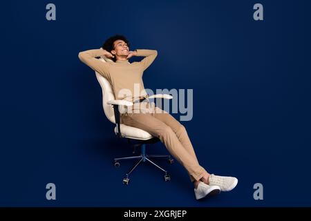 Photo pleine longueur de beau jeune homme assis fauteuil Sleepy ont le repos habillé élégant vêtement beige isolé sur fond de couleur bleu foncé Banque D'Images