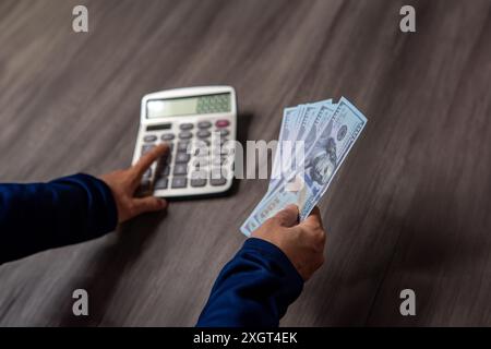 Photo en gros plan des mains d'une personne à l'aide d'une calculatrice et tenant un tas d'argent sur une table en bois. Concept financier. Banque D'Images