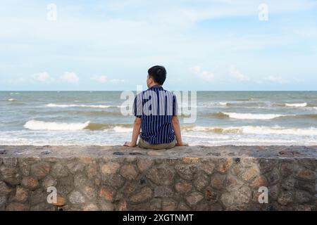 Vue arrière d'un jeune homme asiatique assis sur la promenade dans la mer tropicale le week-end d'été Banque D'Images