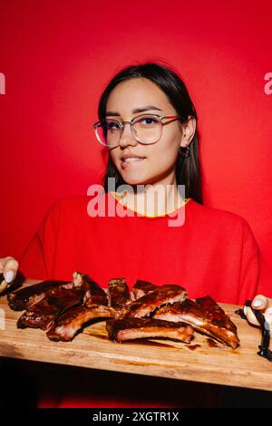 Une jeune femme dans un pull rouge vif sourit tout en tenant une planche de bois chargée de succulentes côtes de barbecue sur un fond rouge vif Banque D'Images