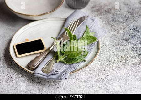 Une charmante table pour l'été avec des assiettes en céramique blanche accentuées de feuilles de menthe fraîche, argenterie vintage et une petite étiquette de tableau noir, le tout Banque D'Images