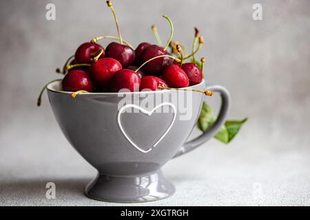 Cerises rouges mûres dans un bol gris moderne avec une découpe en forme de coeur, placé sur un fond gris texturé. L'image capture la couleur éclatante et la FRE Banque D'Images