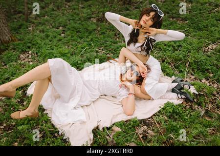 Deux amies profitant d'un moment de détente dans les bois tout en étant allongées sur une couverture habillée de blanc et stylisée avec des lunettes de soleil Banque D'Images