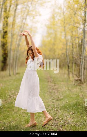 Une jeune femme joyeuse aux cheveux roux bouclés, portant une robe blanche, danse au milieu de grands arbres minces dans une forêt luxuriante, exprimant la liberté et le bonheur dedans Banque D'Images