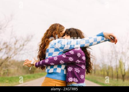 Deux femmes, portant des pulls colorés à motifs, partagent une étreinte chaleureuse dans une zone boisée, célébrant leur amitié au milieu d'un paysage naturel apaisant Banque D'Images