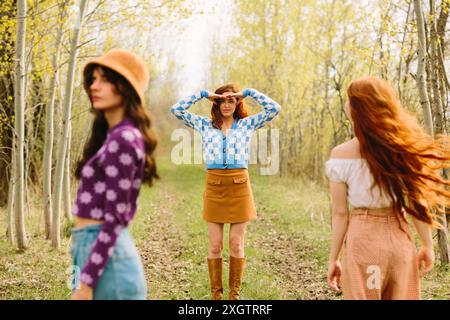 Trois femmes élégantes marchent à travers une forêt ensoleillée, entourées d'arbres en herbe. La figure centrale, portant un pull à carreaux bleu et blanc, se tient debout Banque D'Images