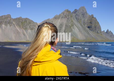 Portrait d'une femme voyageuse blonde en veste jaune debout regardant le paysage Vestrahorn à Stokksnes Banque D'Images