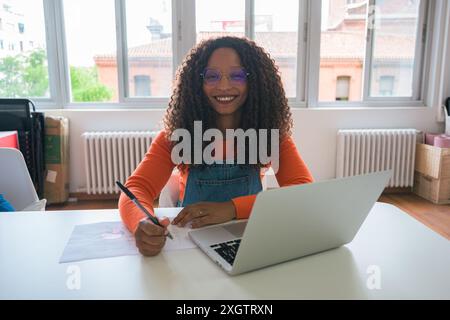 Une femme noire aux cheveux bouclés sourit tout en travaillant sur un ordinateur portable elle est assise à un bureau blanc, portant une chemise orange vif et une salopette bleue, dans un WE Banque D'Images