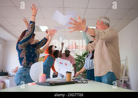 Un groupe diversifié de collègues jetant joyeusement des papiers dans l'air, célébrant le succès dans leur environnement de bureau lumineux, transmettant un sentiment de Banque D'Images