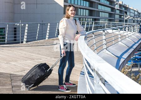 Une jeune femme voyageuse marche tranquillement avec une valise sur un pont urbain moderne, profitant du soleil chaud et d'un ciel dégagé Banque D'Images