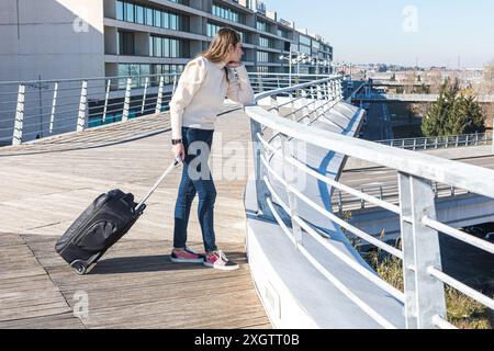 Une jeune femme avec une valise regarde loin en se tenant debout sur un pont urbain moderne, signalant un moment de voyage ou de transition Banque D'Images