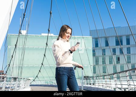 Une femme vêtue de façon décontractée, tenant ses bagages avec un smartphone à la main, regarde son téléphone portable tout en se tenant debout sur un pont ensoleillé avec des bâtisses modernes Banque D'Images