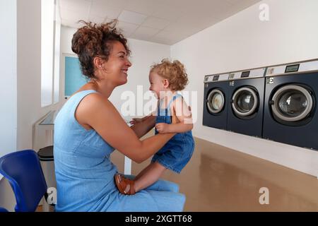 Femme hispanique jouant avec son fils en attendant la fin de la lessive à la laverie automatique. Laverie en libre-service. Banque D'Images