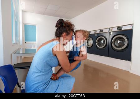 Femme hispanique jouant avec son fils en attendant la fin de la lessive à la laverie automatique. Laverie en libre-service. Banque D'Images