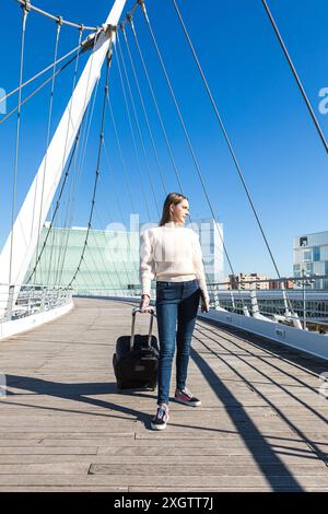 Une jeune femme tirant une valise traverse un pont à haubans moderne par une journée ensoleillée, avec des bâtiments en arrière-plan elle regarde loin de t Banque D'Images