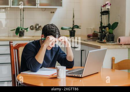 Un jeune homme adulte est assis à une table de cuisine, visiblement stressé avec ses mains sur son front alors qu'il travaille sur un ordinateur portable Banque D'Images