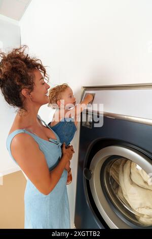 Enfant en bas âge insérant une pièce de monnaie dans la fente de la machine à laver. Femme hispanique mettant la pièce de monnaie dans la machine à laver pour fonctionner. Laverie en libre-service Banque D'Images