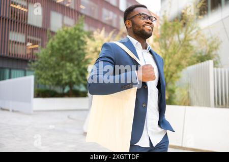 Homme d'affaires afro-américain dans un costume élégant marchant en toute confiance à l'extérieur, regardant loin de la caméra, et souriant. Banque D'Images