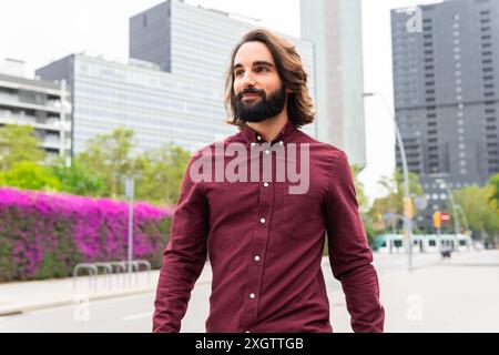 Un jeune homme élégant avec une barbe marche en toute confiance dans une rue bordée d'arbres à Poblenou, Barcelone, vêtu d'une élégante chemise Bordeaux, appréciant le Banque D'Images