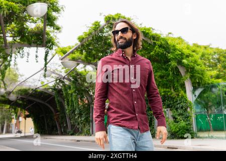 Un homme élégant en lunettes de soleil et chemise marron marche en toute confiance par un pont couvert de lierre à Poblenou, reflétant un style de vie urbain mélangé avec décontracté f Banque D'Images