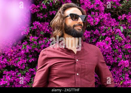 Un homme barbu à l'allure tendance se tient en toute confiance dans une chemise Bordeaux décontractée complétée par des lunettes de soleil à la mode, sur un fond éclatant de p. Banque D'Images