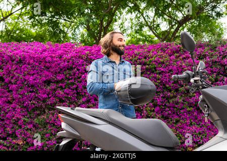 Un homme joyeux dans une veste en Jean tient un casque à côté de sa moto, avec un fond de fleurs violettes luxuriantes, capturant une scène de style de vie dynamique dans Banque D'Images