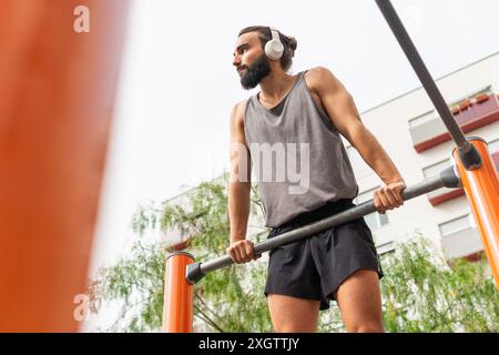 Angle bas de jeune homme concentré avec une barbe s'entraîne dans un parc de fitness en plein air à Poblenou, écoutant de la musique à travers son casque, enhancin Banque D'Images