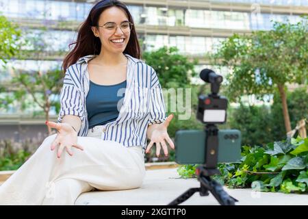 Une femme joyeuse en tenue de printemps décontractée se filme avec un smartphone posé sur un trépied, assise à l'extérieur entourée de verdure Banque D'Images