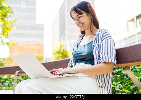 Une jeune femme joyeuse avec des lunettes et une chemise rayée sourit tout en travaillant sur son ordinateur portable, assise sur un banc de parc, entourée de verdure luxuriante dans un an Banque D'Images