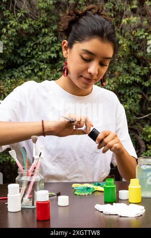 Une jeune femme concentrée aime peindre sur l'artisanat en argile à l'extérieur, en utilisant des couleurs acryliques vives et divers pinceaux, entourés d'une toile de fond naturelle Banque D'Images