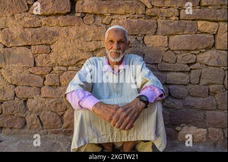 Cette image capture un homme marocain âgé dans une tenue traditionnelle, assis avec confiance contre un mur de pierre rustique son expression et sa posture stable p Banque D'Images