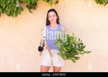 Une jeune femme se tient contre un mur, tenant dans une main des cisailles à élaguer et dans l'autre des branches vertes fraîchement coupées. Elle est habillée en gardenin décontracté Banque D'Images