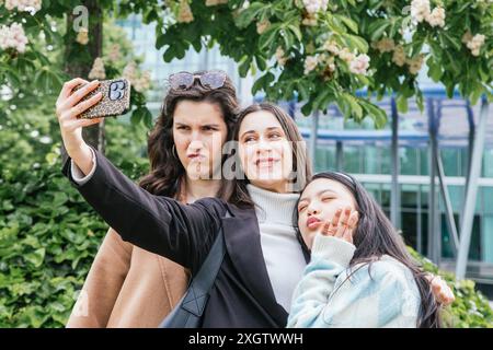 Trois jeunes amies posant pour un selfie en plein air, chacune affichant une expression ludique unique dans un cadre de parc florissant Banque D'Images