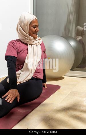 Une femme arabe souriante dans un hijab fait du yoga sur un tapis à l'intérieur, avec un grand ballon d'exercice en arrière-plan. Elle regardait loin rayonnant de paix et de con Banque D'Images