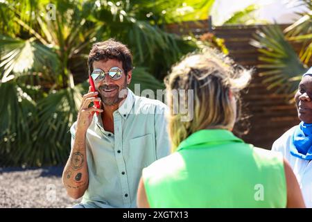Des collègues d'affaires multiraciaux discutent à l'extérieur dans un cadre tropical. Un homme caucasien parle au téléphone, souriant, portant des lunettes de soleil, détournant le regard. A Banque D'Images