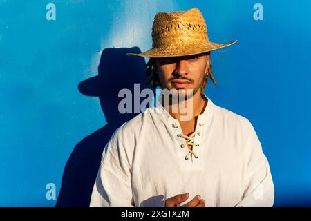 Un jeune homme caucasien avec des dreadlocks et un chapeau de paille se tient sur un fond bleu vif. Il regarde directement la caméra avec un expr confiant Banque D'Images