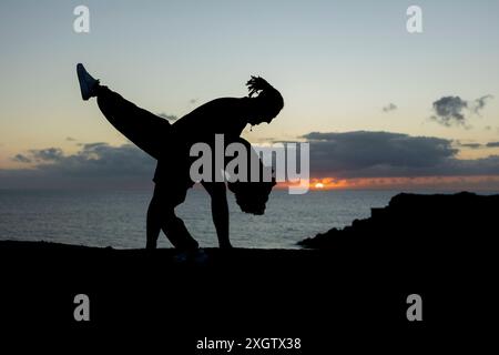 Un homme avec des dreadlocks effectue un mouvement de danse acrobatique en silhouette contre un coucher de soleil vibrant sur la mer, se penchant en arrière dans une pose dynamique. Banque D'Images