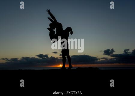 Un homme et une femme méconnaissables exécutent un mouvement de danse dynamique en silhouette contre un coucher de soleil vibrant sur l'océan, capturant un moment de gracieuse intera Banque D'Images