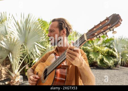 Un homme aux cheveux longs joue de la guitare à l'extérieur, entouré de plantes tropicales luxuriantes. Il porte une chemise dorée et regarde loin de la caméra. Banque D'Images